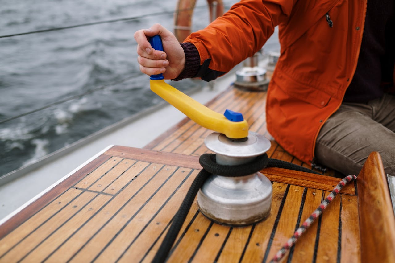 services-03 Man in orange jacket operating winch on sailboat deck during daytime sailing adventure.