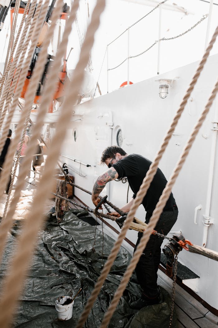 services-01 A tattooed sailor manages ropes on a well-bound ship under sunlight, showcasing seamanship.